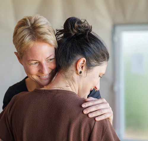 Zwei Frauen umarmen sich herzlich in einem hellen Raum, strahlende Gesichter und Nähe zeigen emotionale Verbundenheit.