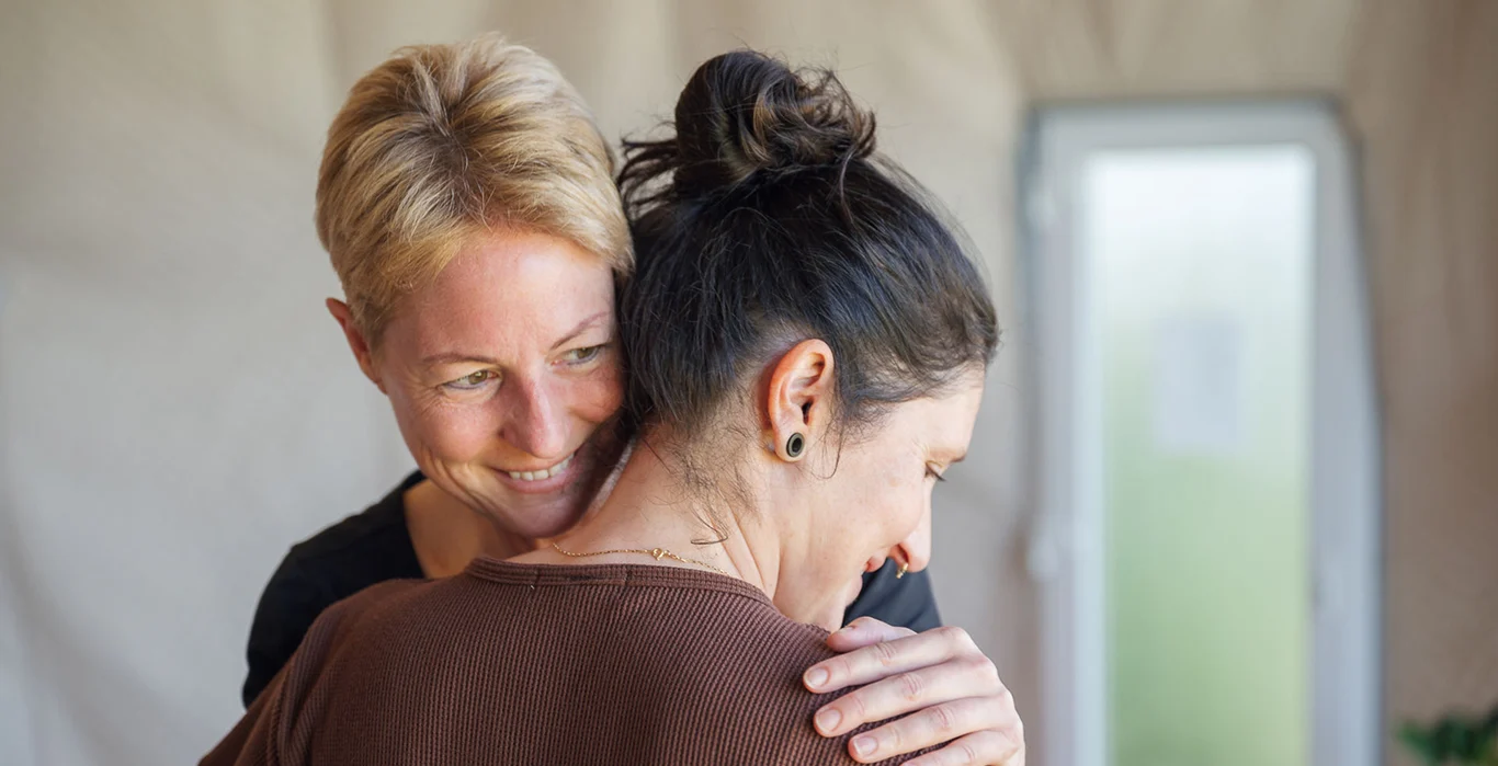 Zwei Frauen umarmen sich liebevoll in einem hellen Raum, strahlende Gesichter und emotionale Verbundenheit.