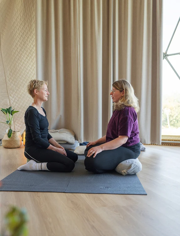 Zwei Frauen sitzen sich im Yoga-Studio gegenüber und praktizieren Achtsamkeit und Meditation auf einer Matte.